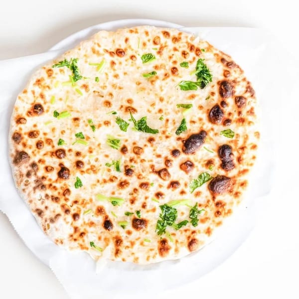 Round Indian naan bread, golden brown with charred spots and fresh green herbs on a white plate.